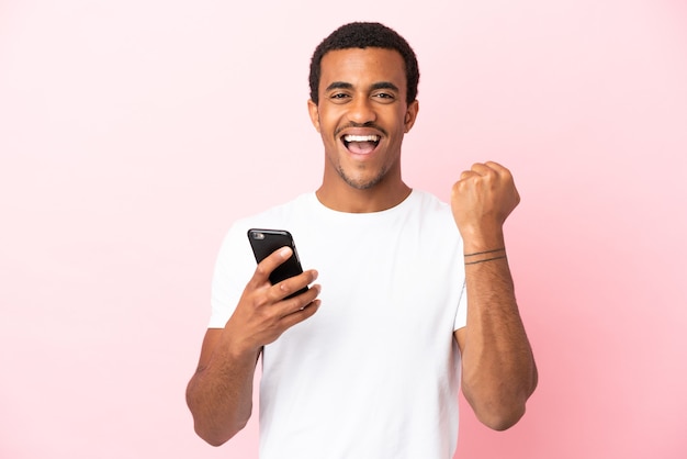 Homem afro-americano bonito em fundo rosa isolado usando telefone celular e fazendo gesto de vitória