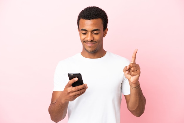 Homem afro-americano bonito em fundo rosa isolado usando telefone celular e levantando o dedo