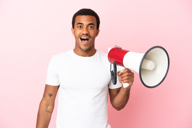 Homem afro-americano bonito em um fundo rosa isolado segurando um megafone e com expressão de surpresa