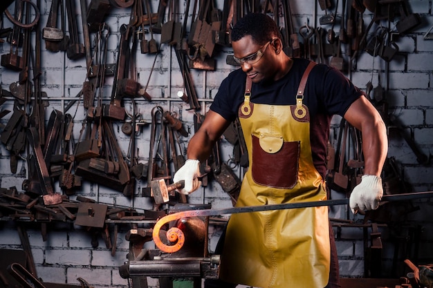 Homem afro-americano bonito forjando aço ao lado do forno na oficina escura. conceito de pequenas empresas.