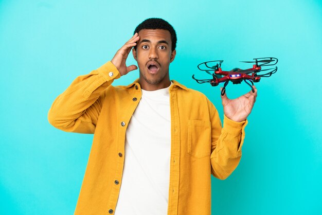 Homem afro-americano bonito segurando um drone sobre um fundo azul isolado com expressão de surpresa