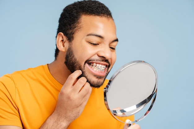 Homem afro-americano com aparelhos ortodônticos examinando seus dentes enquanto olha em um espelho