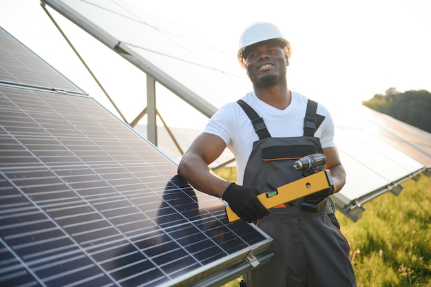 Homem afro-americano com capacete branco e macacão cinza entre fileiras de painéis solares