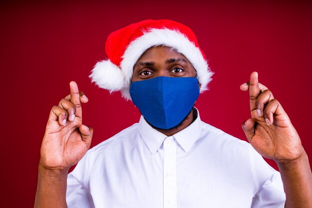 Homem afro-americano com chapéu de papai noel usando máscara em fundo de estúdio vermelho