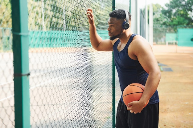 Homem afro-americano com fones de ouvido sem fio faz uma pausa e se apoia na malha de metal com bola na quadra ao ar livre