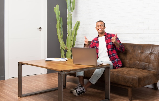 Homem afro-americano com laptop na sala de estar dando um polegar para cima gesto com as duas mãos e sorrindo