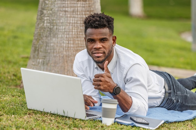 Homem afro-americano com thumbsup deitado no chão enquanto usa um computador portátil