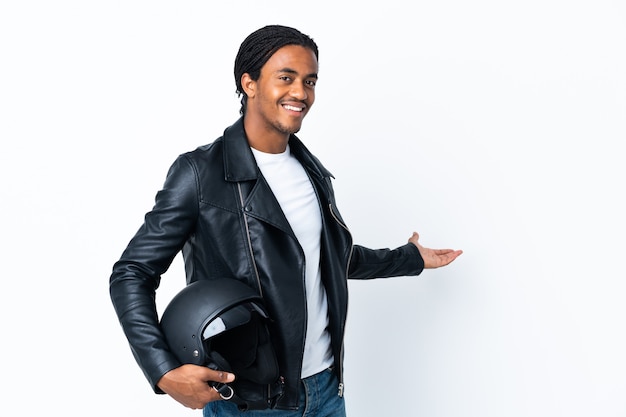 Homem afro-americano com tranças, segurando um capacete de motociclista isolado na parede branca, estendendo as mãos para o lado para convidar a vir