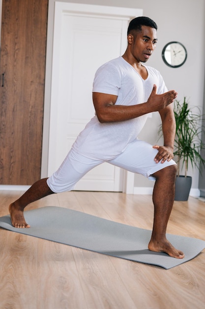 Homem afro-americano de ajuste focado fazendo exercício de fitness esportivo em pé no tapete de ioga em uma sala doméstica brilhante, olhando para longe. Conceito de treinamento esportivo no ginásio em casa.