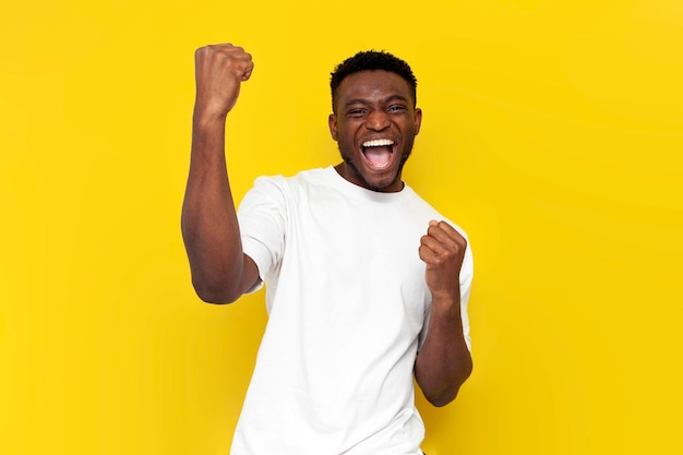 Homem afro-americano de camisa branca celebra a vitória e levanta as mãos em fundo amarelo