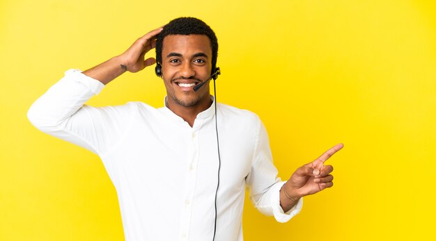 Homem afro-americano de telemarketing trabalhando com um fone de ouvido sobre um fundo amarelo isolado surpreso e apontando o dedo para o lado