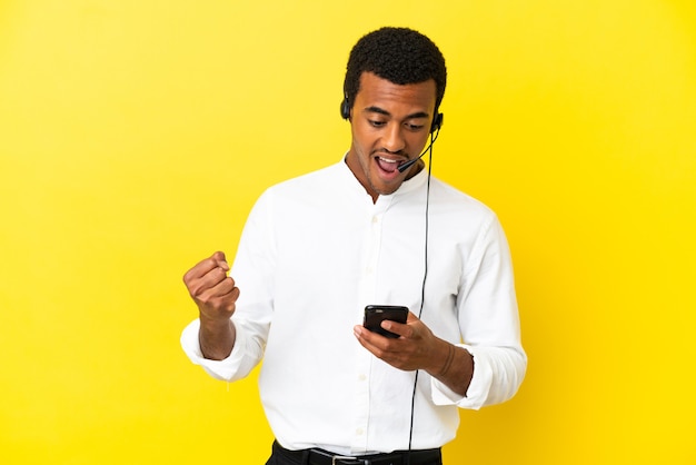 Homem afro-americano de telemarketing trabalhando com um fone de ouvido sobre um fundo amarelo isolado, surpreso e enviando uma mensagem