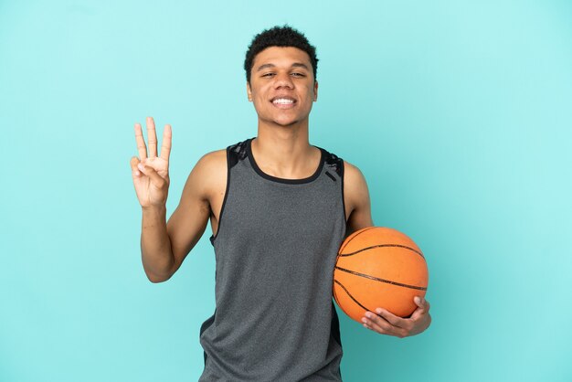 Homem afro-americano do jogador de basquete isolado em um fundo azul feliz e contando três com os dedos