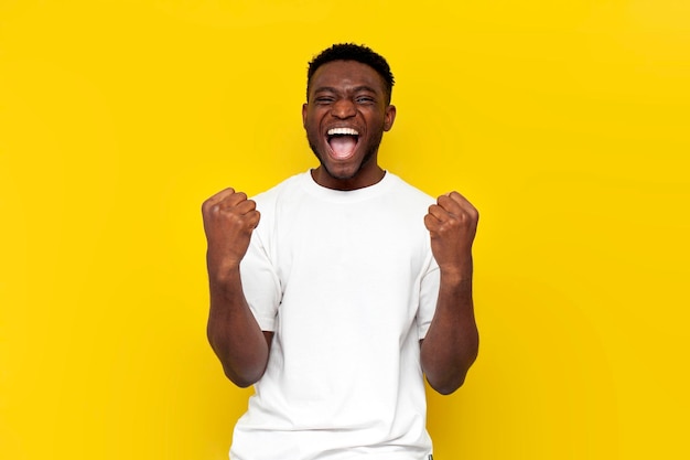 Homem afro-americano em camiseta branca comemora a vitória e levanta as mãos sobre fundo amarelo