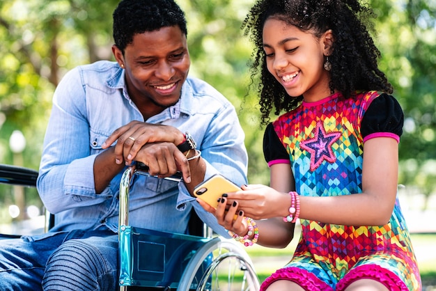 Homem afro-americano em uma cadeira de rodas, usando um telefone celular com a filha, enquanto desfruta de um dia juntos no parque.