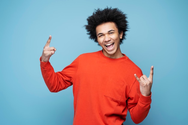 Homem afro-americano emocional gritando, mostrando sinal de rocha isolado em fundo azul