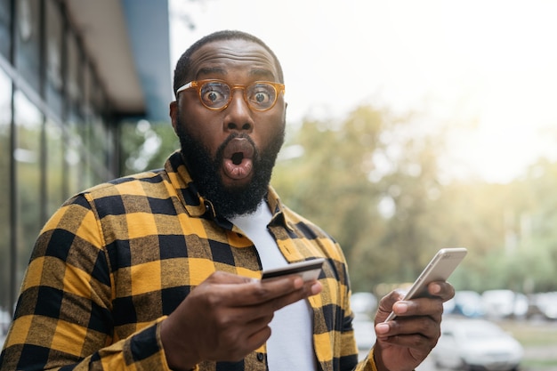 Homem afro-americano emocional, segurando um cartão de crédito, compras online. Freelancer emocional recebe pagamento
