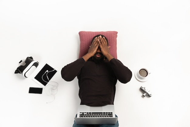 Homem afro-americano emocional usando laptop rodeado por gadgets isolados na parede branca do estúdio.