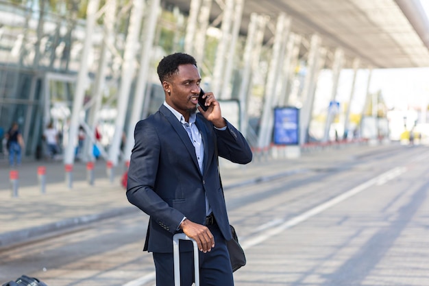 Homem afro-americano falando ao celular perto do aeroporto, empresário chegou em viagem de negócios