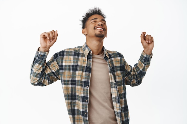 Homem afro-americano feliz dançando e curtindo a festa, levantando as mãos com expressão facial despreocupada, regozijando-se, dançando e se divertindo, de pé contra um fundo branco