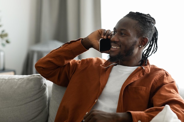 Homem afro-americano feliz falando no celular em casa sorrindo cara fazendo ou atendendo chamadas por telefone sentado no sofá