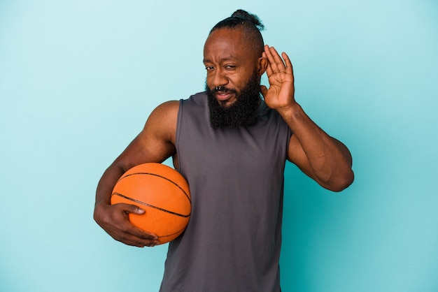 Homem afro-americano jogando basquete isolado na parede azul, tentando ouvir uma fofoca.