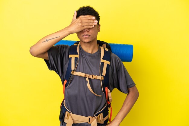 Homem afro-americano jovem alpinista com uma grande mochila isolada em um fundo amarelo, cobrindo os olhos com as mãos. Não quero ver nada