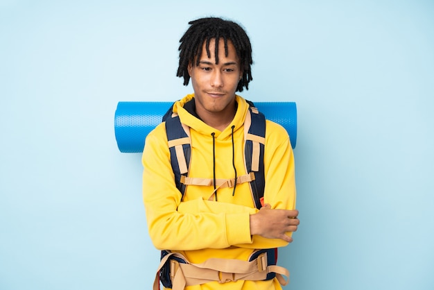 Homem afro-americano jovem alpinista com uma mochila grande em uma parede azul, pensando em uma idéia