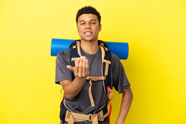 Homem afro-americano jovem alpinista com uma mochila grande isolada em um fundo amarelo, convidando para vir com a mão. Feliz que você veio