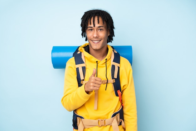 Foto homem afro-americano jovem alpinista com uma mochila grande isolada em uma parede azul, dando um polegar para cima gesto