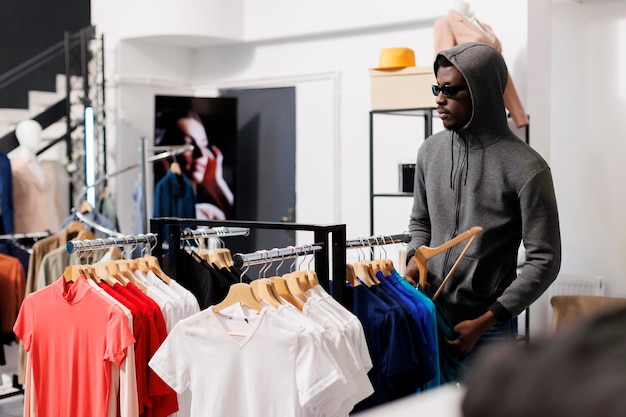 Homem afro-americano ladrão tomando um risco, olhando para roupas de moda antes de roubá-lo em uma boutique moderna. Ladrão usando óculos de sol e capuz não ser reconhecido no centro comercial. Conceito de assalto