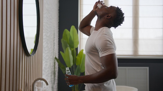 homem afro-americano limpando dentes enxaguar com produto de cuidados dentários higiene bucal no banheiro enxaguar