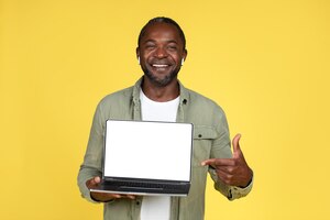 Homem afro-americano maduro sorridente em fones de ouvido sem fio casuais mostra o dedo no laptop com tela em branco