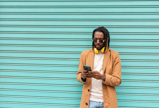 Homem afro-americano moderno com óculos de sol elegantes, enviando uma mensagem com o celular Isolado em fundo azul