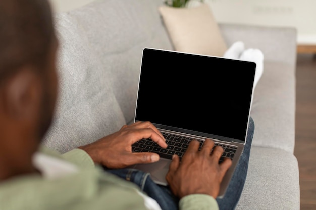 Homem afro-americano ocupado de meia-idade no sofá assiste a vídeo digitando no laptop com tela em branco nas horas vagas