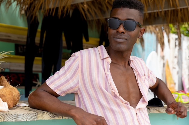 Homem afro-americano relaxado em óculos de sol, apoiando-se no balcão da barraca de praia de aluguel de surf. Negócio local, hobbies, desporto, surf, verão e férias, inalterados.