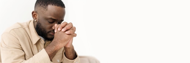 Homem afro-americano rezando pela paz enquanto está sentado no sofá em casa Banner da Web