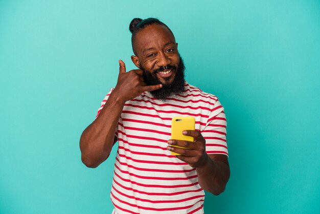 Homem afro-americano, segurando um telefone celular isolado em um fundo azul, mostrando um gesto de chamada de celular com os dedos.