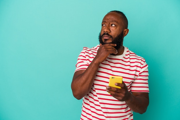 homem afro-americano segurando um telefone celular isolado em um fundo azul, olhando de soslaio