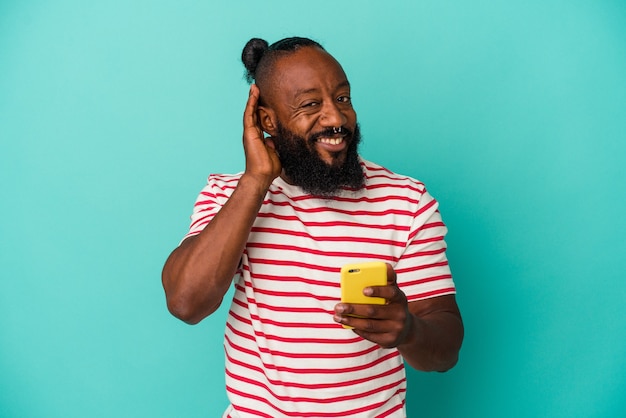 Homem afro-americano, segurando um telefone móvel isolado em um fundo azul, tentando ouvir uma fofoca.