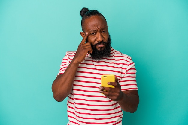 Homem afro-americano, segurando um telefone móvel isolado no fundo azul, apontando o templo com o dedo, pensando, focado em uma tarefa.