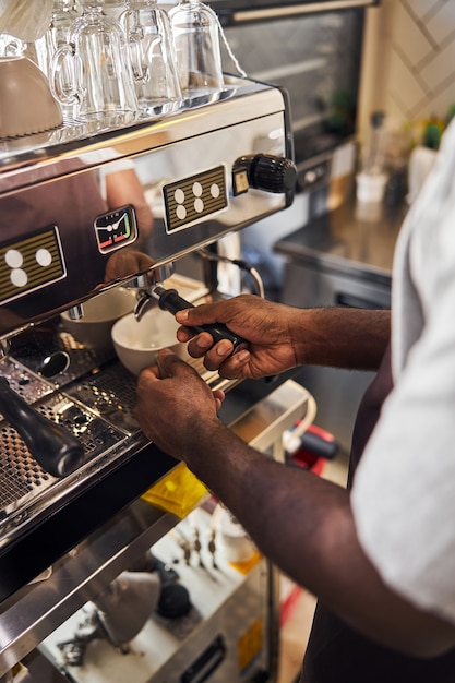 Homem afro-americano segurando uma xícara branca sob a torneira da máquina de café enquanto prepara um expresso fresco