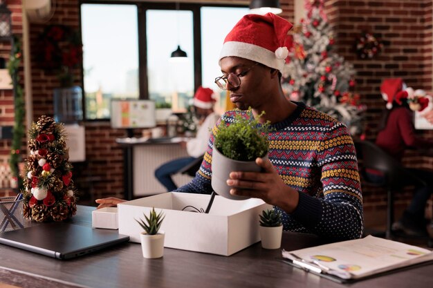 Homem afro-americano sendo demitido na véspera de natal, reunindo pertences para deixar o emprego no escritório com decorações festivas de natal. Sentindo-se triste e chateado com o fracasso na carreira e o emprego perdido.