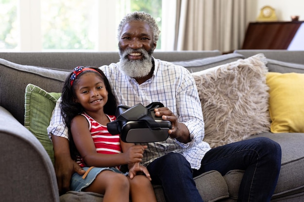 Homem afro-americano sênior e sua neta aproveitando seu tempo juntos em casa, sentados em um sofá, segurando um fone de ouvido VR, olhando para a câmera e sorrindo