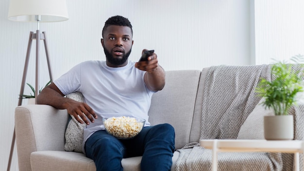 Homem afro-americano senta-se em um sofá em uma sala de estar vendo televisão ele está segurando um controle remoto