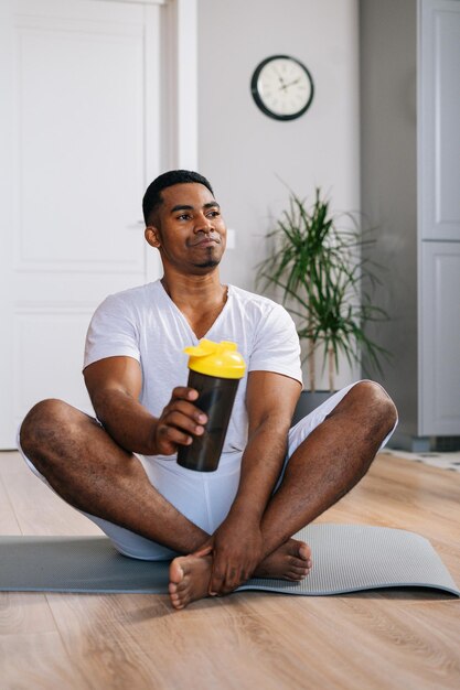 Homem afro-americano sentado de pernas cruzadas e relaxando no colchonete após treino água potável