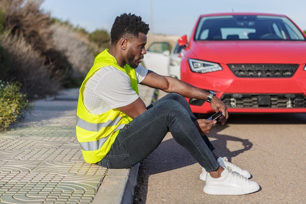 Homem afro-americano sentado em uma calçada ao lado de um carro vermelho quebrado