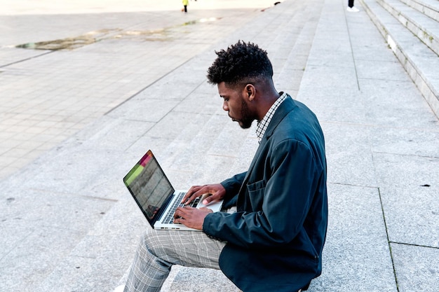 Homem afro-americano sentado nas escadas da cidade de costas usando seu laptop para trabalho ou videoconferência com uma tela em branco