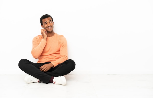 Foto homem afro-americano sentado no chão sobre uma parede isolada de copyspace, pensando em uma ideia enquanto olha para cima