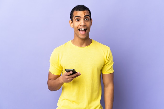 Homem afro-americano sobre parede roxa isolada usando telefone celular com expressão facial de surpresa